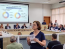 Audiência pública sobre o orçamento de São Carlos no plenário da Câmara Municipal, com telão exibindo receitas e despesas municipais e cidadã segurando o Guia do Cidadão
