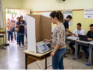 Eleitora vota em urna eletrônica durante eleição municipal em seção de votação, com mesários e fila de eleitores ao fundo